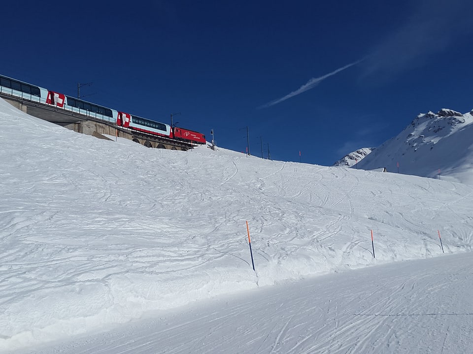 Der Glacier Express fährt durch die verschneite Landschaft mit blauem Himmel.