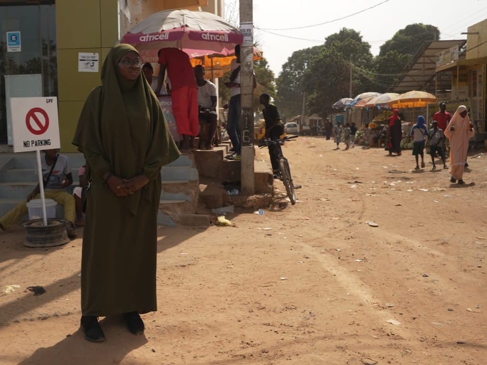 Fatoumatta steht an der Kreuzung. Im Hintergrund sieht man viele Menschen, die den Strassenmarkt besuchen.