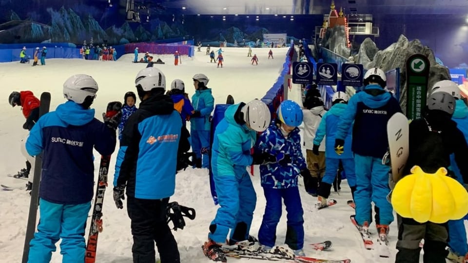 Menschen stehen an einem Förderband an um wieder ans obere Ende der Ski-Piste zu gelangen. 