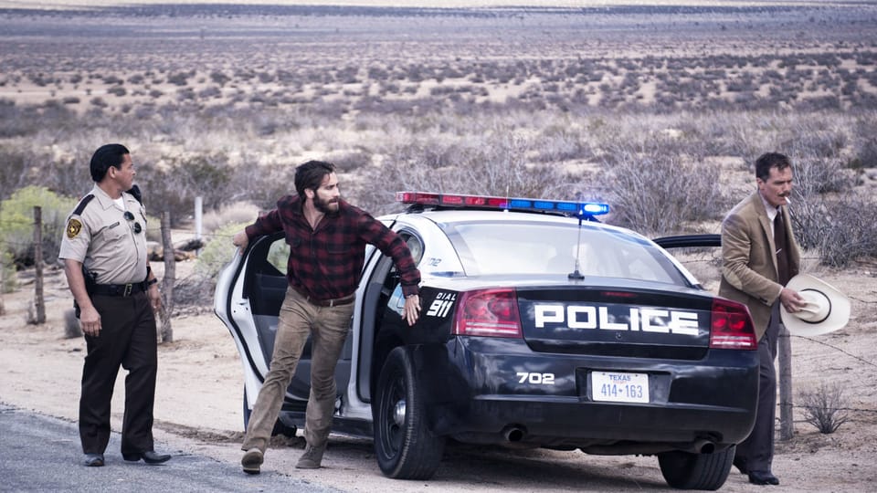 Ein Polizist und zwei Männer in Zivil stehen neben einem Polizeiauto in der Wüste.