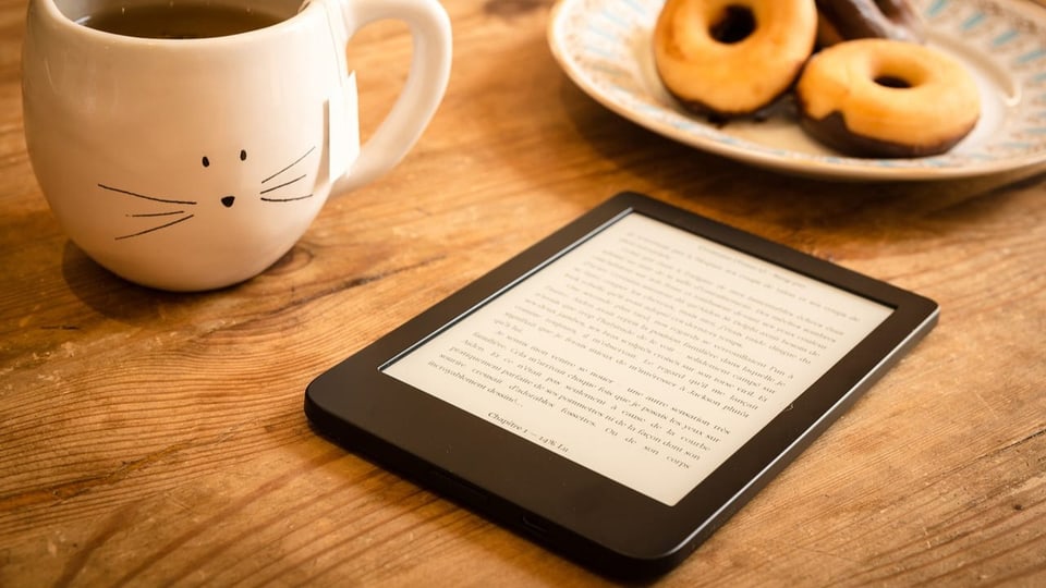 Ein E-Reader liegt auf dem Tisch zusammen mit einer Tasse Tee und Mini-Donuts.