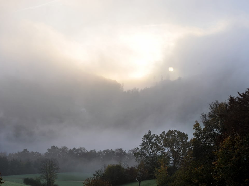 Foto in Graustufen. Wäler und Hügel mit Nebelschwaden, teils drückt die Sonne durch. Auch die Scheibe der Sonne ist zu erkennen. 