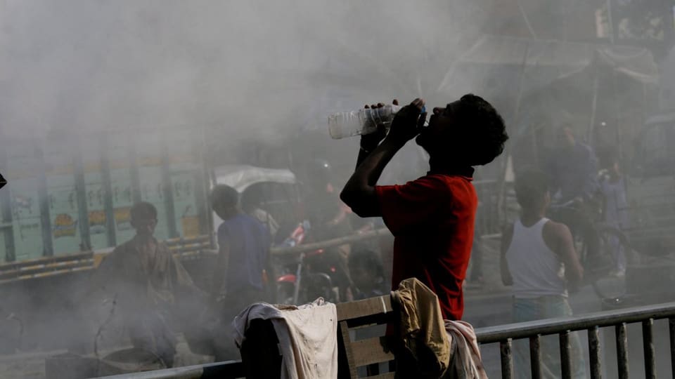 Ein Mann trinkt Wasser in Kalkutta, Rauch steigt hinter ihm auf 