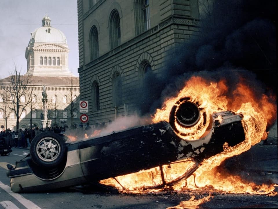 Ein Auto liegt auf dem Dach und brennt – im Hintergrund das Bundeshaus.