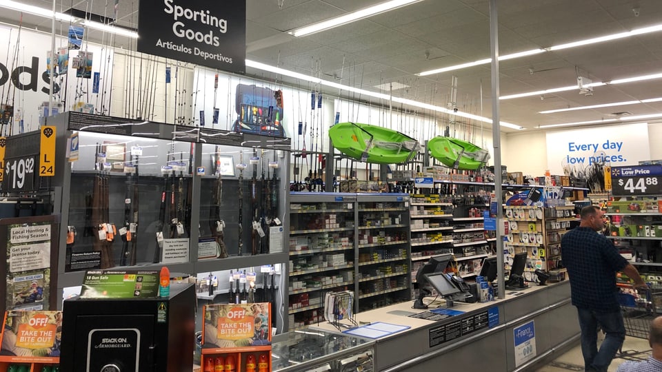 Sportabteilung in einem Supermarkt. Man sieht die oben erwähnten Gegenstände. Eine Vitrine mit Gewehren. Ein Mann mit Einkaufswagen läuft daran vorbei.