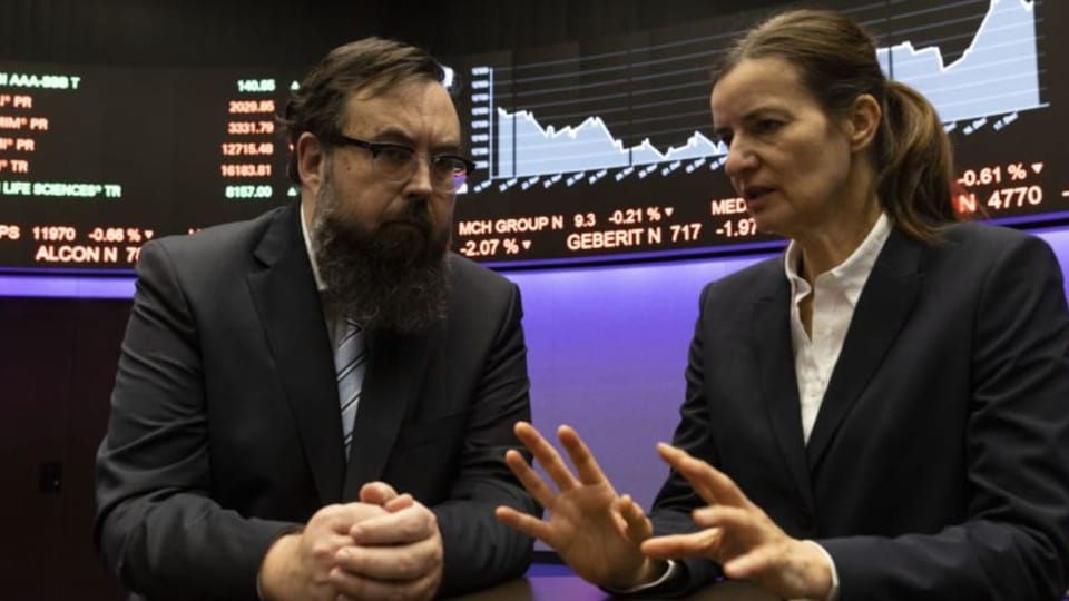 Ein Mann und eine Frau vor einem Screen, auf dem Börsenkurse zu sehen sind.