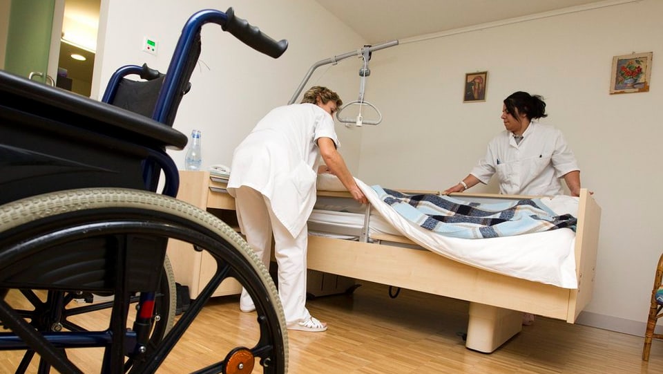 Zwei Pflegerinnen machen ein Bett. Im Vordergrund steht ein Rollstuhl.