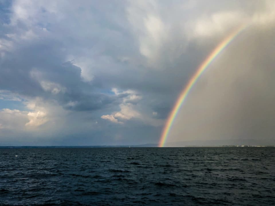 Rechts im Bild ein Regenbogen, der bis zum Wasser reicht. Man blickt auf den See hinaus. Der Himmel ist bewölkt. 