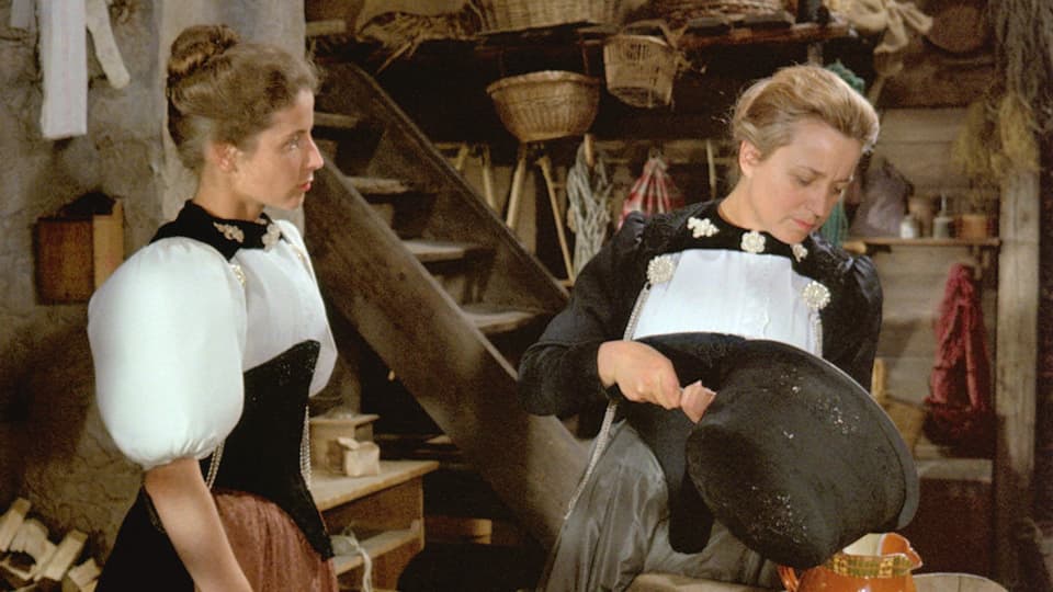 Zwei Frauen in Tracht stehen in einer Bauernküche. Diejenige zur Rechten giesst gerade eine Tee in eine Kanne.