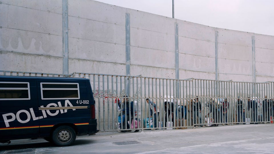 Eine Mauer, vor der ein Polizeiauto steht und Menschen mit Tüten