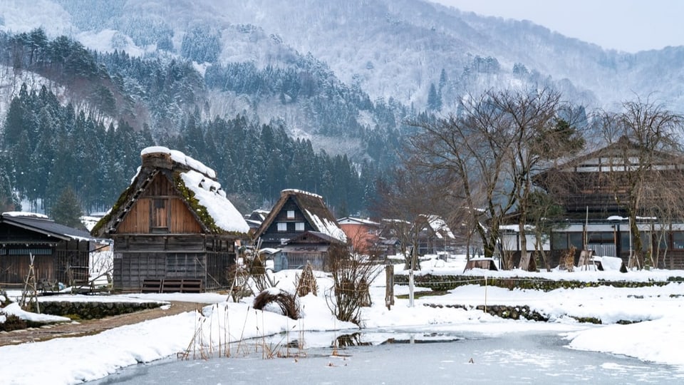 Das Bild zeigt verschneite Holzhäuser und davor einen gefrorerenen Teich.