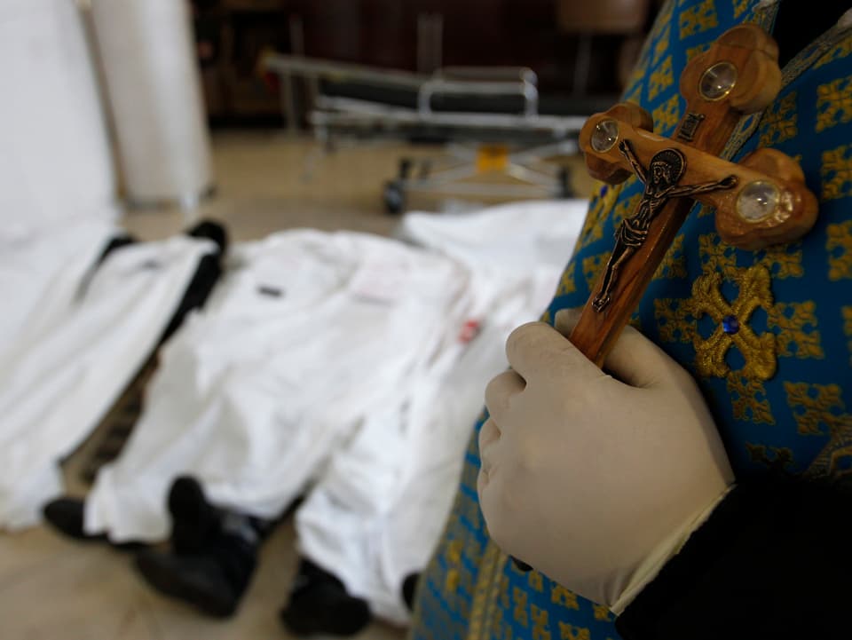 Ein Priester hält ein Kreuz in der linken Hand, im Hintergrund liegen mehrere Leichen unter weissen Tüchern.