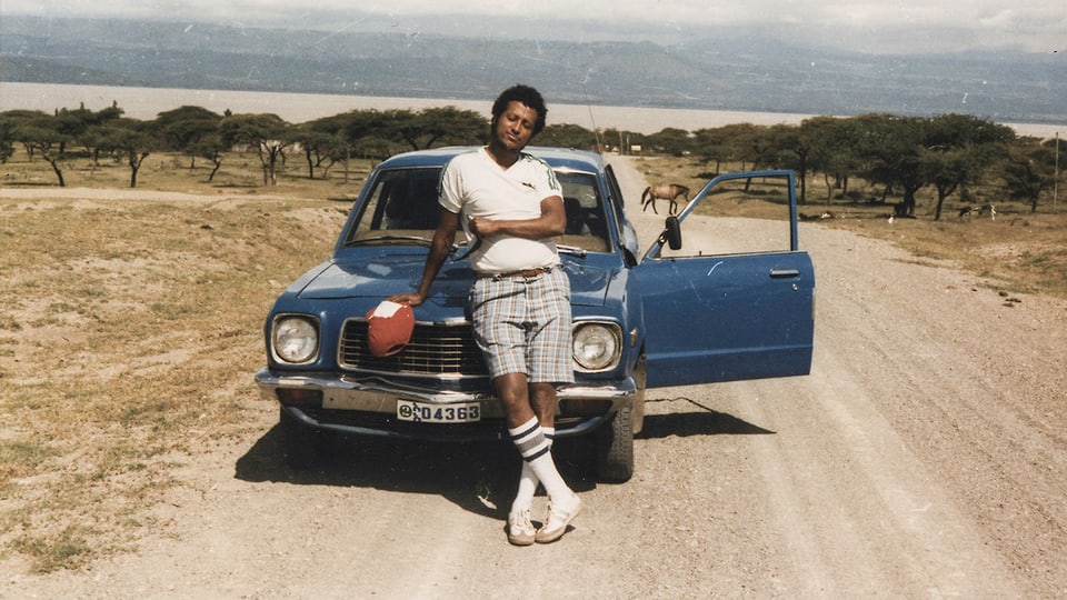 Ein junger schwarzer Mann in weissen Tennissocken vor einem blauen Auto.