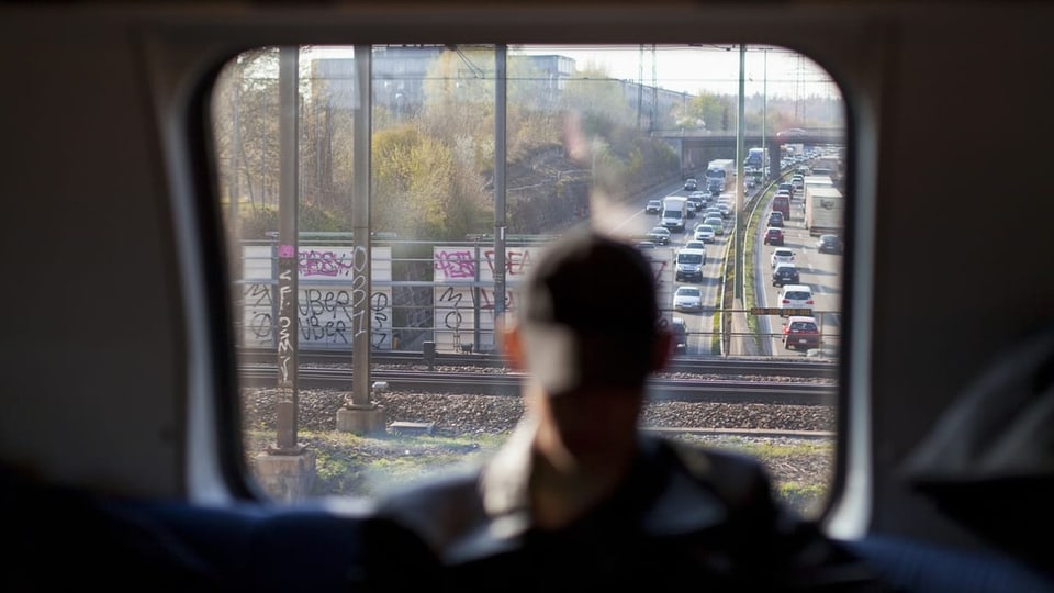 A man in front of a train window, cars behind him on a highway in a traffic jam.