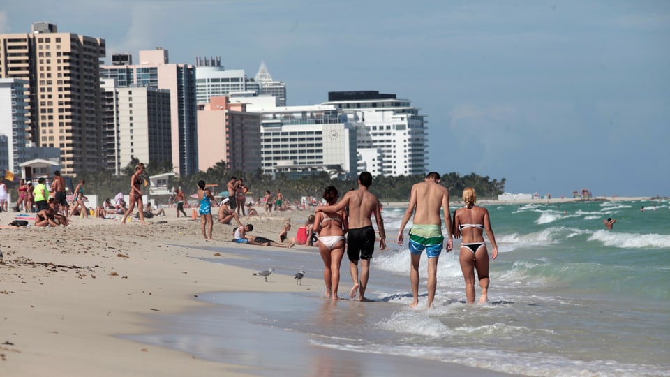 Menschen laufen in Badekleidern am Strand von Miami Beach, im Hintergrund Hochhäuser.