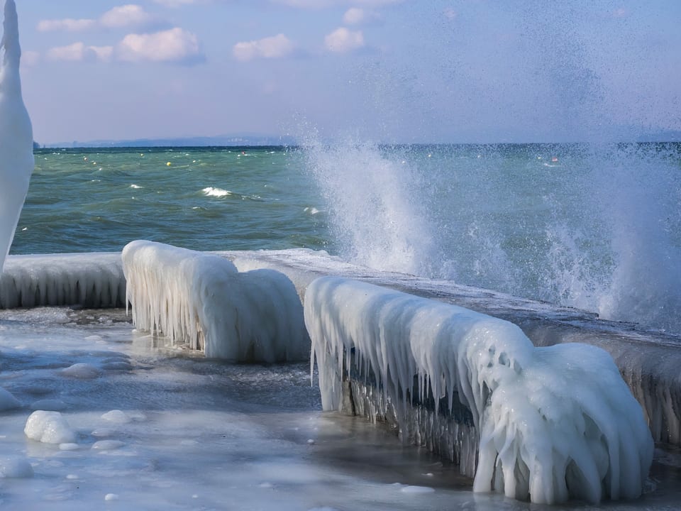 Uferpromenade mit Sitzbank, die von Eis überzogen ist. Auf dem Wasser mit Bise Wellen. 
