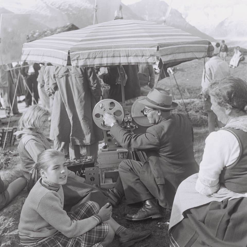 Die Kamera steht unter einem Schirm auf der Alp und wird von einem Mann gewartet. Neben ihm sitzen zwei Mädchen und eine Frau, die ihm zuschauen.