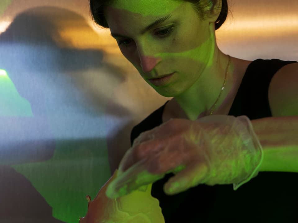 Eine Frau wird von verschiedenen Lichtern beleuchtet. Mit einem Gummihandschug hält sie ein undefinierbares Objekt in der Hand.