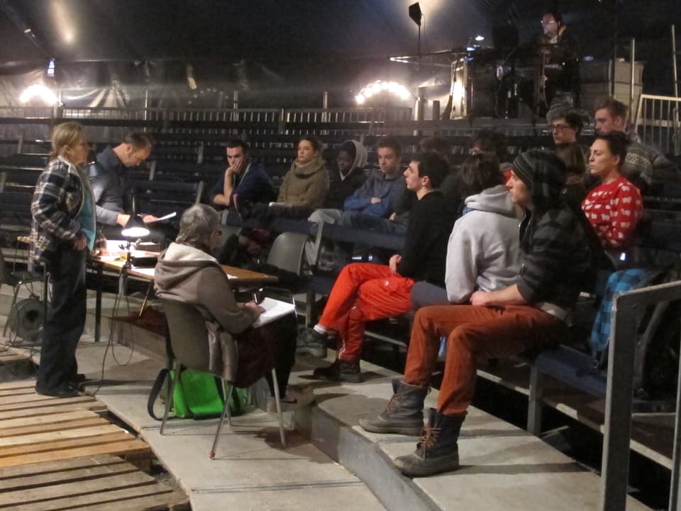 Mehrere Leute sitzen auf den Zuschauerrängen des Zirkuszeltes und diskutieren.