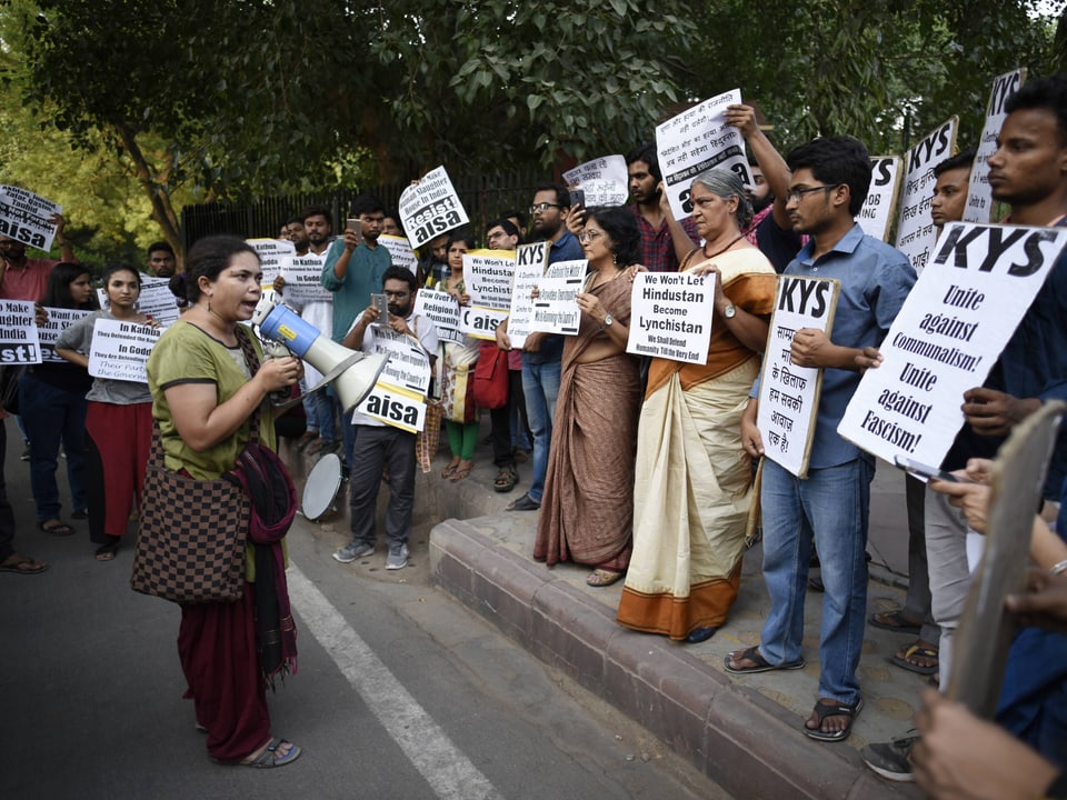 Frau mit Megafon spricht vor Gruppe von Demonstranten mit Plakaten in Indien.