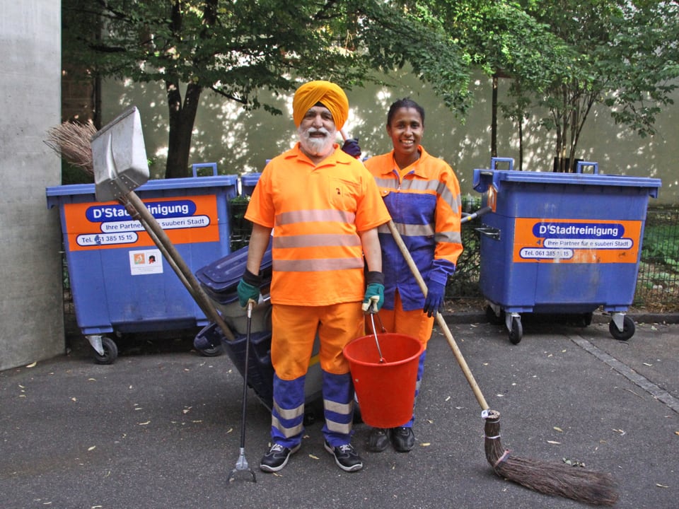 Ein Mann mit orangem Turban steht neben einer Frau. Sie haben Strassenputzsachen dabei.