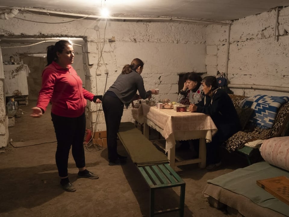 Vier Frauen sind in einem Keller, zwei sitzen, eine steht und gestikuliert mit betrübtem Gesicht.