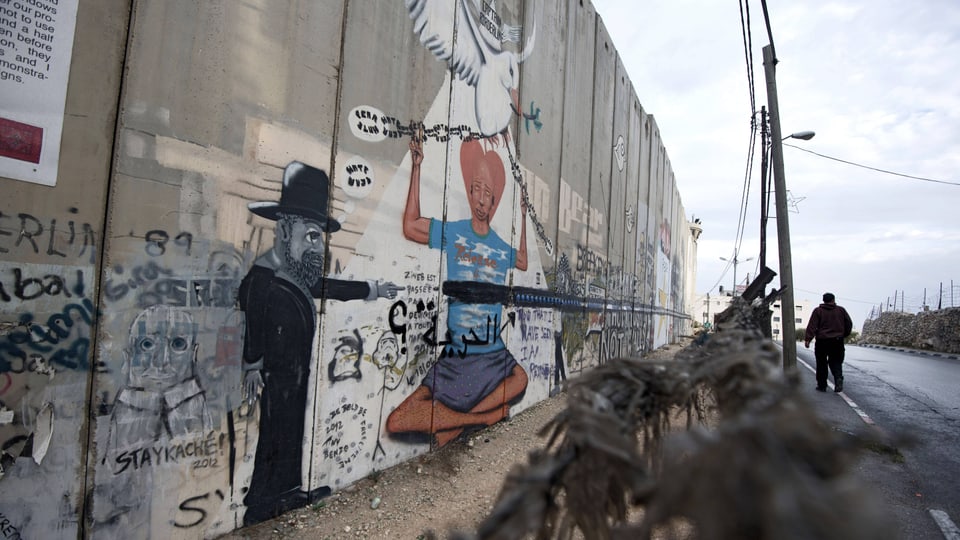 Eine hohe Mauer gesäumt von Stacheldraht, neben einer Strasse. Die Mauer ist mit Street Art und Graffitis bemalt.