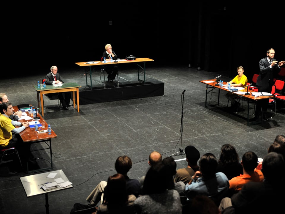 Auf einer Bühne sind Tische so angeordnet wie in einem Gerichtssaal. Das Publikum schaut den Schauspielern zu, die Angeklagten tragen gelbe T-Shirts.