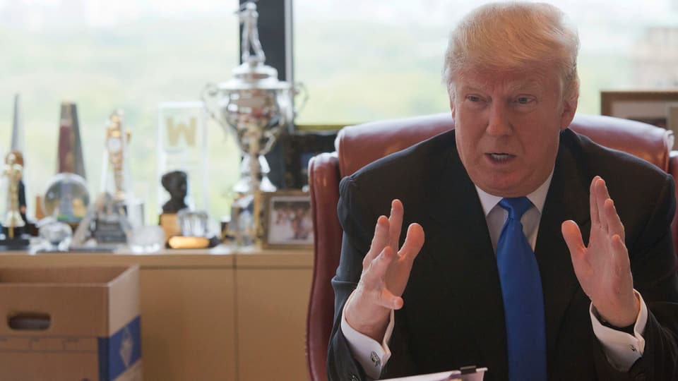 Trump an einem Schreibtisch sitzend und mit den Händen gestikulierend.