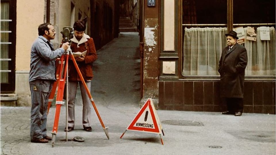 Aufnahme aus den Siebzigerjahren: Zwei Männer vermessen eine Strasse, dahinter Mann mit Hut und Stumpen im Mund