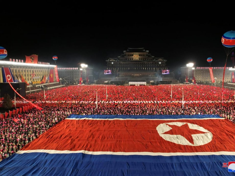 Eine nordkoreanische Flagge breitet sich auf dem Platz aus. Darüber und darunter sind sehr viele Menschen.