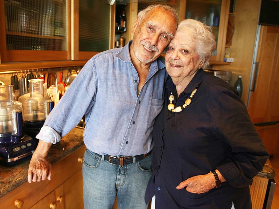 Ein älteres Paar steht in einer Küche – Er hält den Arm um sie.