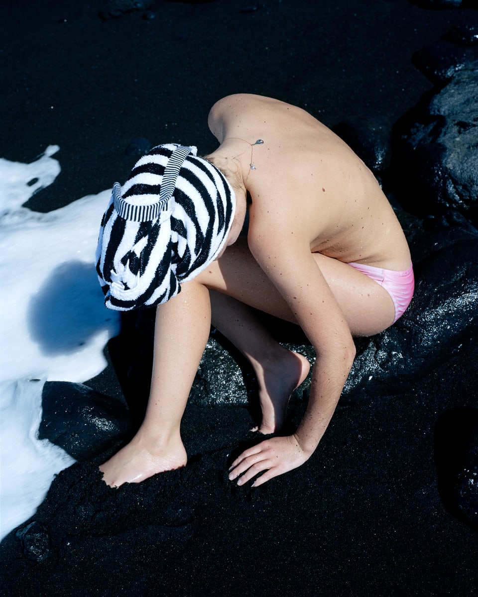 Eine Frau in Unterhose und mit einem Badetuch um den Kopf kauert auf einem schwarzen Sandstrand.
