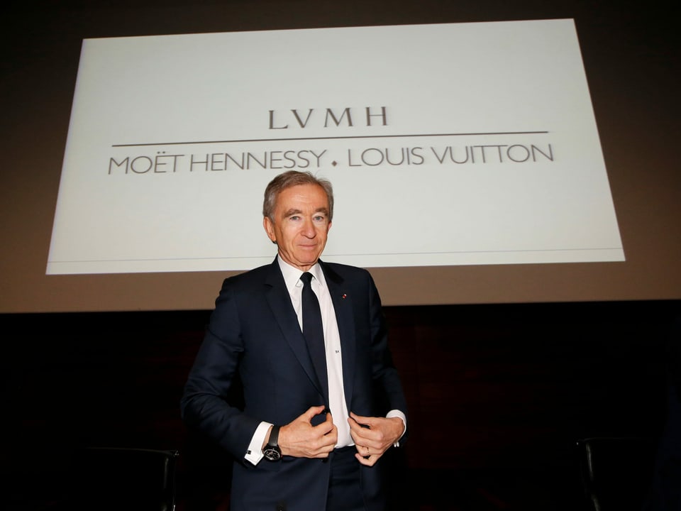 Ein älterer Mann – Bernard Arnault – präsentiert sich vor einem Schild des Luxusgüterkonzerns LVMH. 