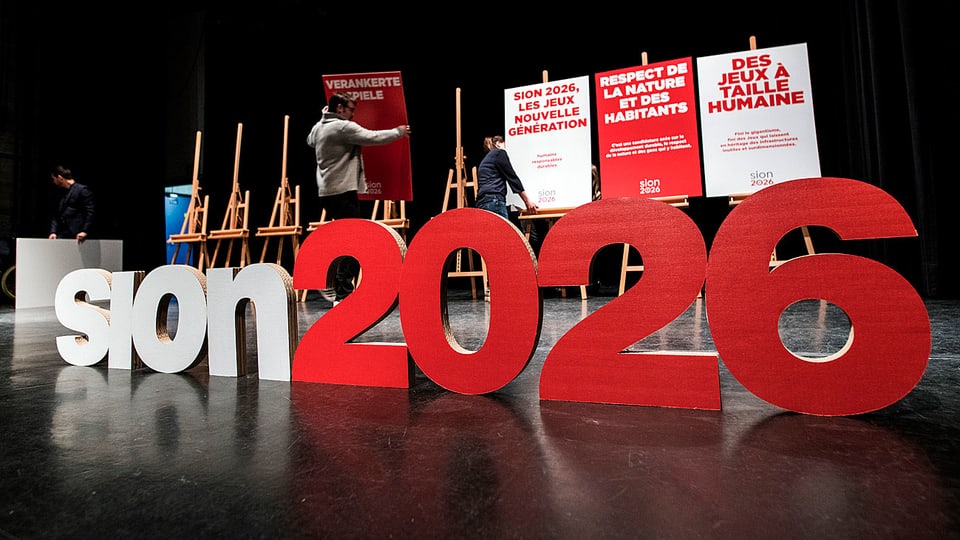 Auf einer Bühne stellen Menschen Plakate auf, im Vordergrund steht in grossen Kartonbuchstaben: Sion 2026.
