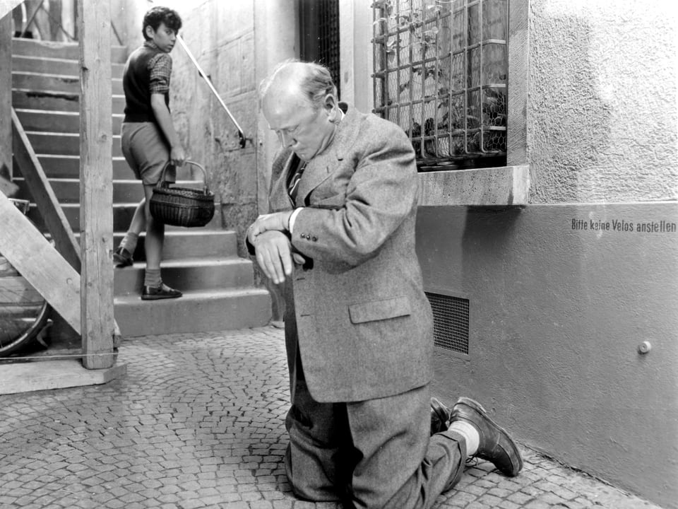 Älterer Mann kniet auf Kopfsteinpflaster und hält sein Handgelenk. Ein Junge mit einem Korb in der Hand steigt eine Treppe hinauf und blickt zum Mann zurück.