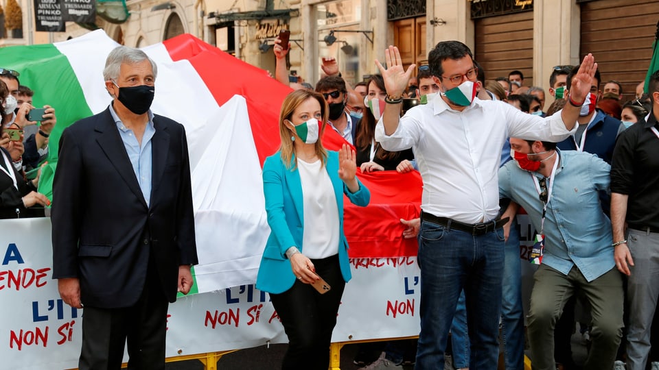 Tajani, Meloni und Salvini mit Mund- und Nasenschutz stehen vor einer italienischen Flagge auf einer Strasse mit anderen Demonstranten.
