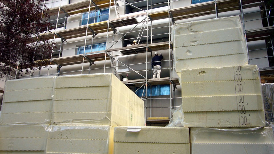 Isolationsmaterial stapelt sich vor einem Wohnblock, an dem die Fassade saniert wird. Bauarbeiter stehen auf dem Gerüst.