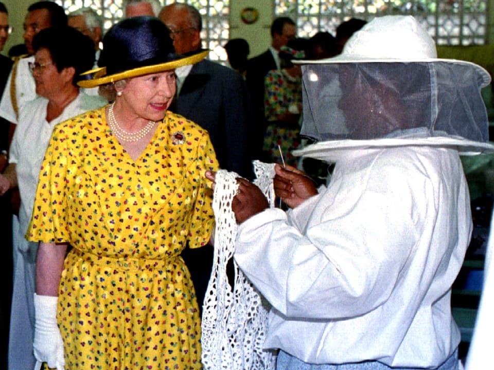 Die Queen spricht mit einer Frau. Diese zeigt ihr ein selbstgenähtes Kleidungsstück.