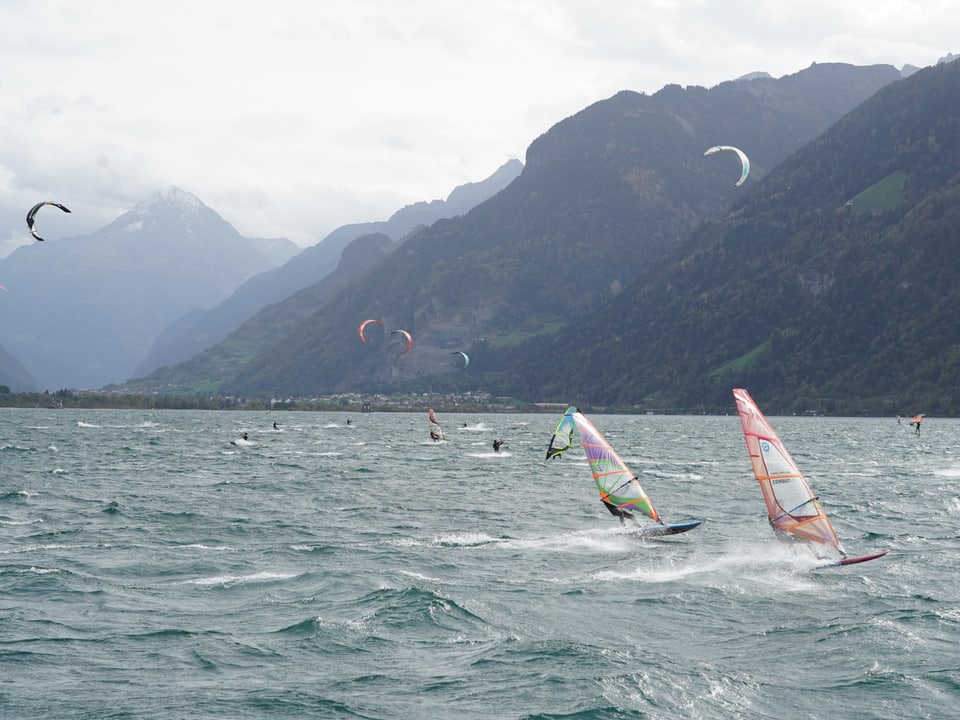 Windsurfer und Kitesurfer auf einem stürmischen See mit Bergen im Hintergrund.