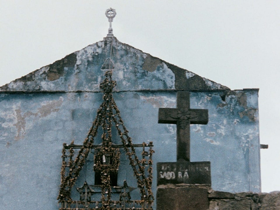 Blick an eine bläuliche Hausfassade mit einem Kreuz im Vordergrund