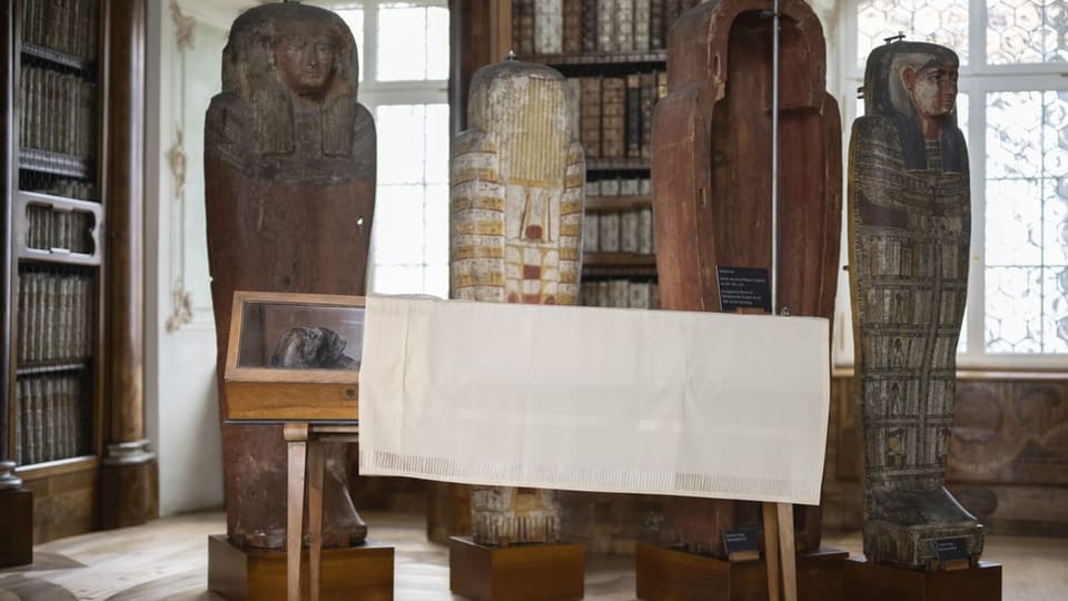 vorne aufgebahrt eine Mumie im gläsernen Sarg, halb bedeckt. Hinten aufgestellt Sarkophage mit Mustern und Hyroglyphen.
