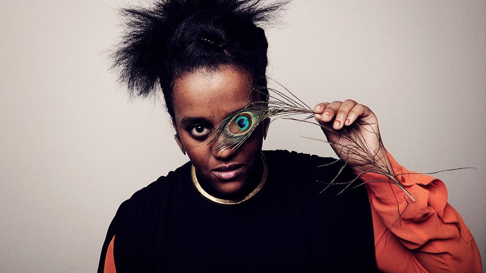 Eine junge Frau hält sich das Auge einer Pfauenfeder vor das eine Auge. 
