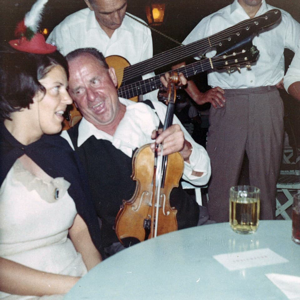 Eine junge Frau sitzt an einem Tisch neben einem Geigenspieler. Hinter ihnen steht ein Mann, der Gitarre spielt.