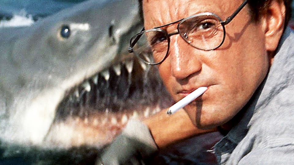 Mann mit Zigarett im Mund schaut in die Kamera. Hinter ihm taucht ein Hai auf.