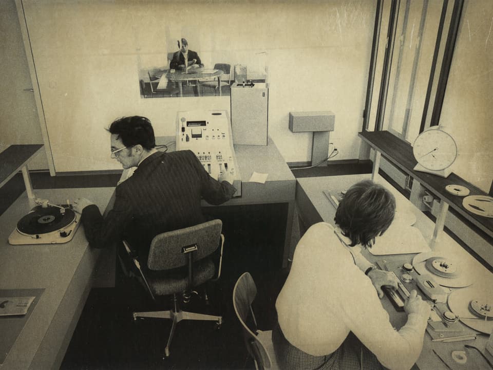 Ein Schwarzweissbild eines Radiostudios: Ein Mann und eine Frau sind an Tonbandmaschinen am Arbeiten.