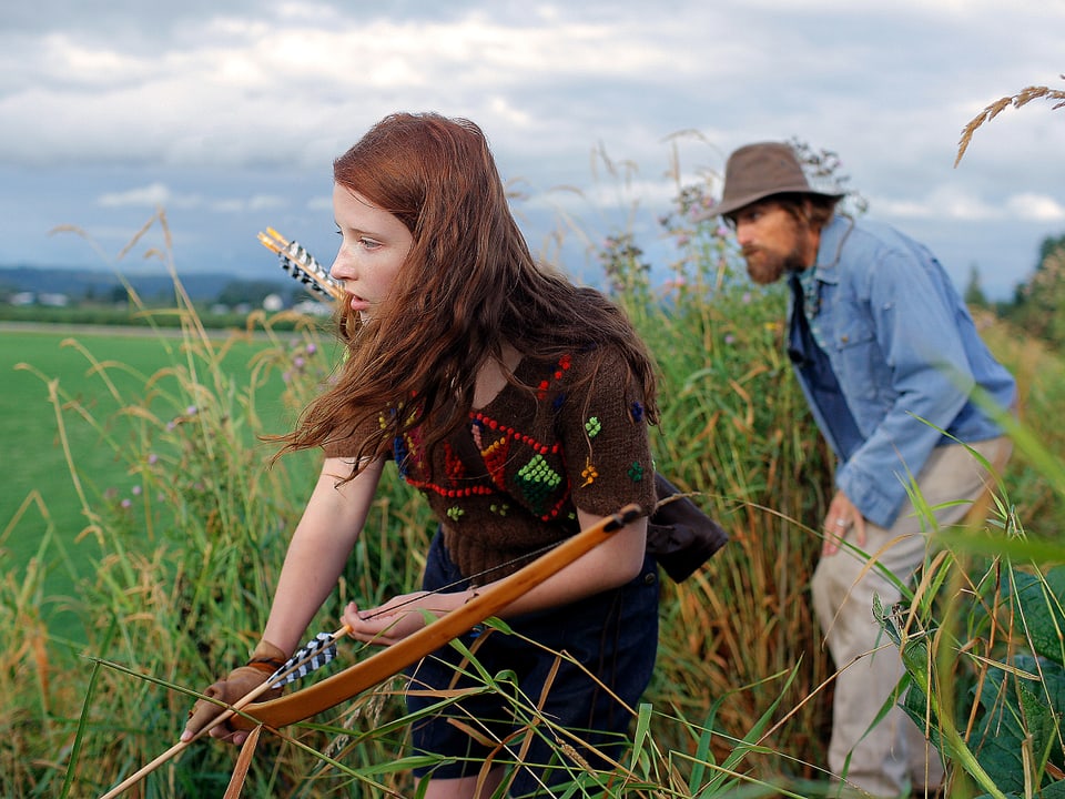 Eine junge Frau mit einem Pfeilbogen auf der Jagd, in ihrem Schlepptau ein Mann mit Jeanshemd und Hut.