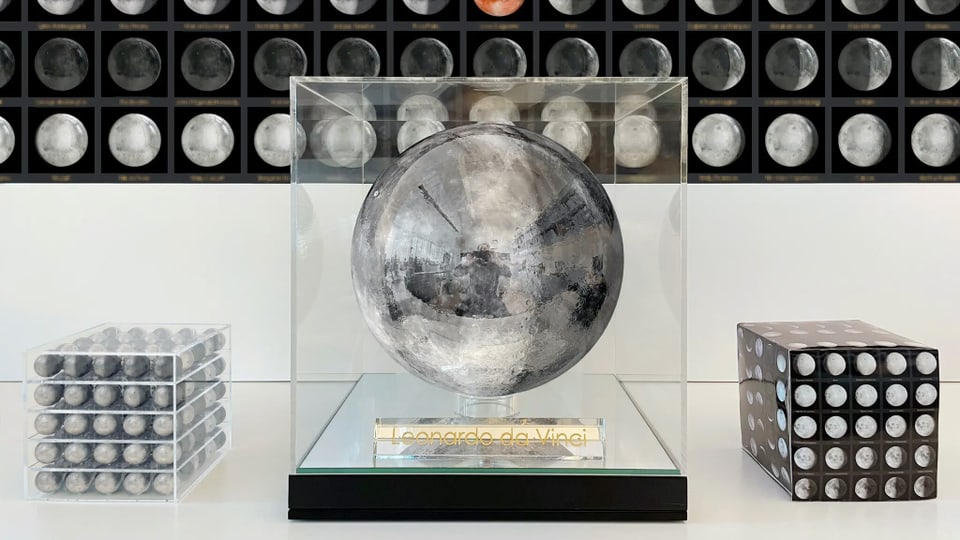 Eine grosse Metallkugel in der Mitte, links in einer Glasbox weitere Metallkugeln. Im Hintergrund sind die Mondphasen.