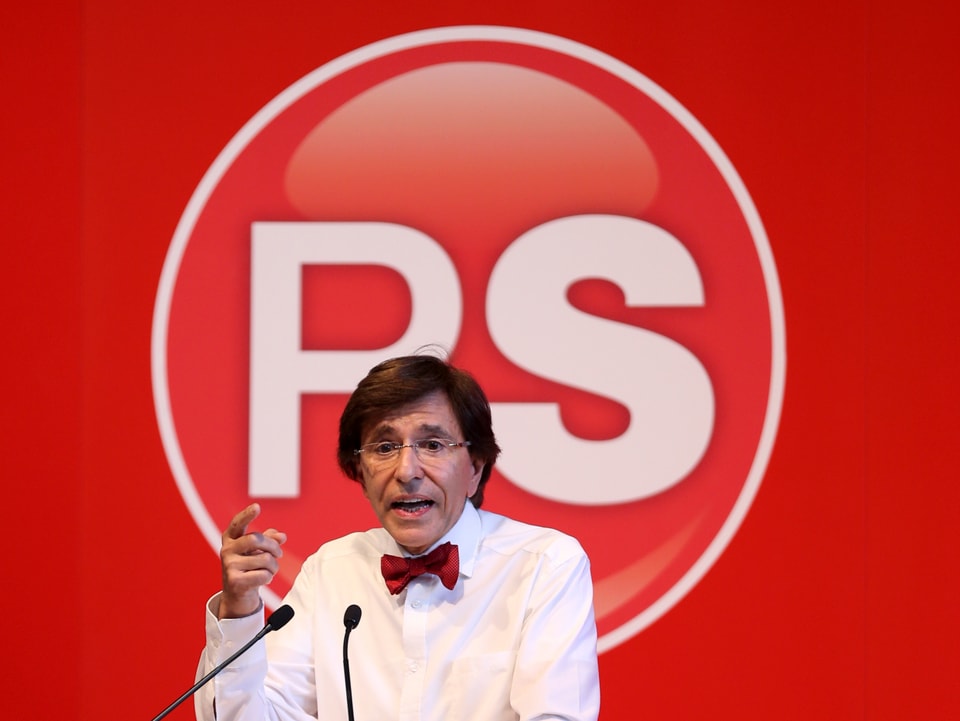 Elio di Rupo mit Fliege vor einer roten Wand mit dem Logo der Parti Socialiste.