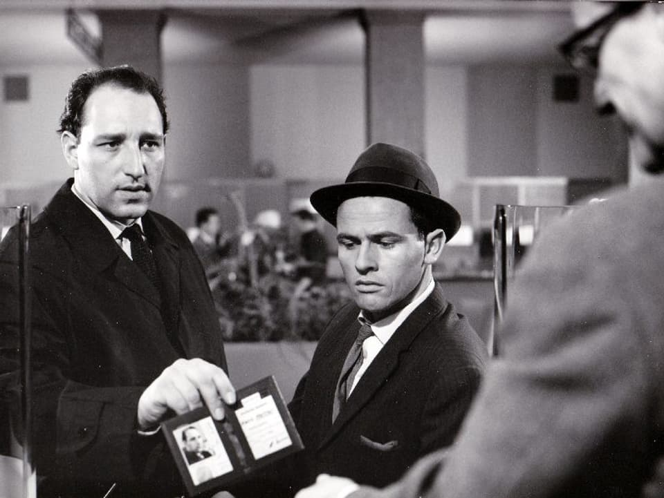 Zwei Männer stehen an einem Schalter. Der grössere der beiden hält dem Mann am Schalter seinen Ausweis entgegen.